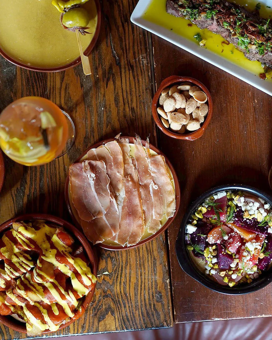 A table of tapas at Perro Negro Taperia from above — jamón over melted cheese, patatas bravas with alioli, marinated beets with pistachio and feta, marcona almonds and cocktails
